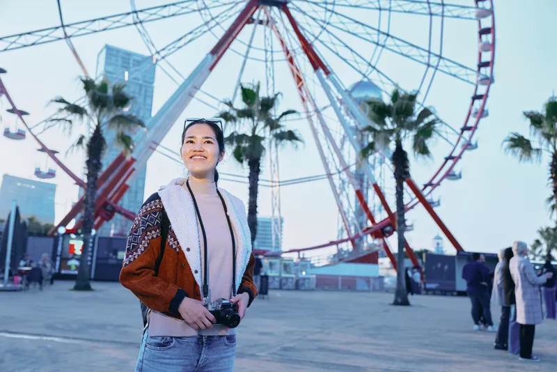 Photography of a traveler holding a camera near a large ferris wheel and palm trees, outdoor urban park scene, casual fashion style, daytime light, candid moment capturing travel and entertainment
