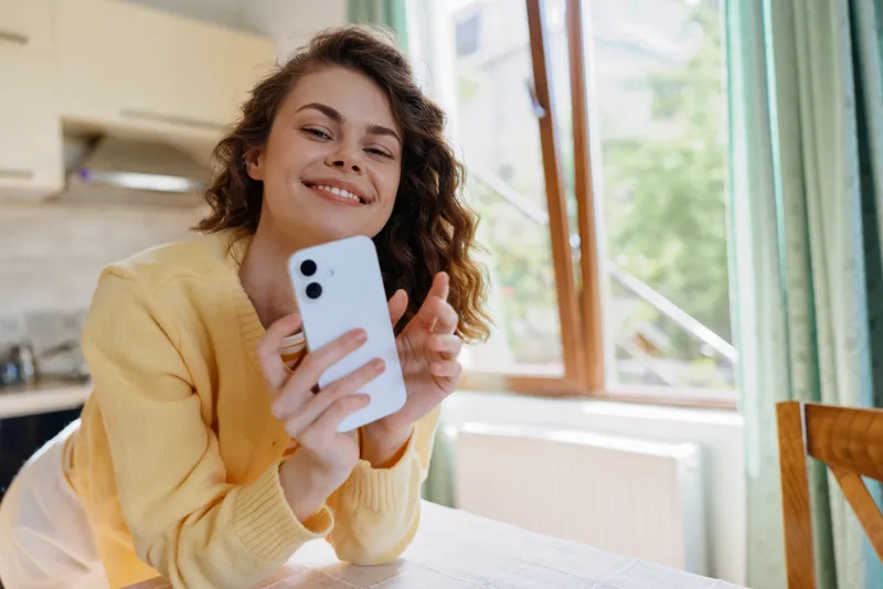 Young woman at home enjoying a moment with a smartphone, candid cheerful expression, casual sweater, bright natural light, cozy kitchen setting, relaxed lifestyle vibe, connected and happy