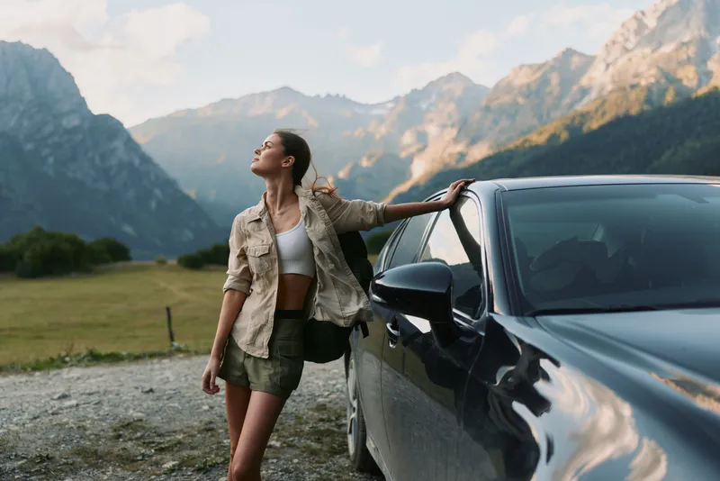A young woman leans against a sleek car in a rugged mountain valley, soaking in fresh air, a sense of freedom and adventure in a wide outdoor landscape.