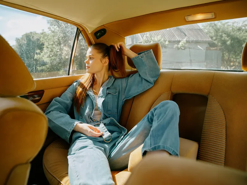 Stylish young woman in a denim outfit, relaxed in a car, enjoying a sunny day. The soft brown interior complements her casual look and thoughtful expression.