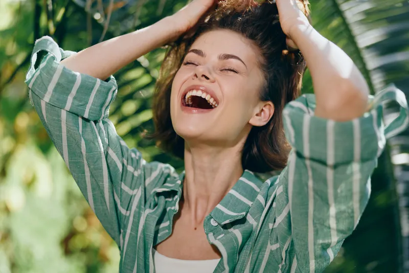 Woman enjoying a bright outdoor moment, smiling with hands in her hair, wearing a striped green shirt, natural lighting, candid portrait, fresh happiness, carefree vibe, soft breeze, vibrant