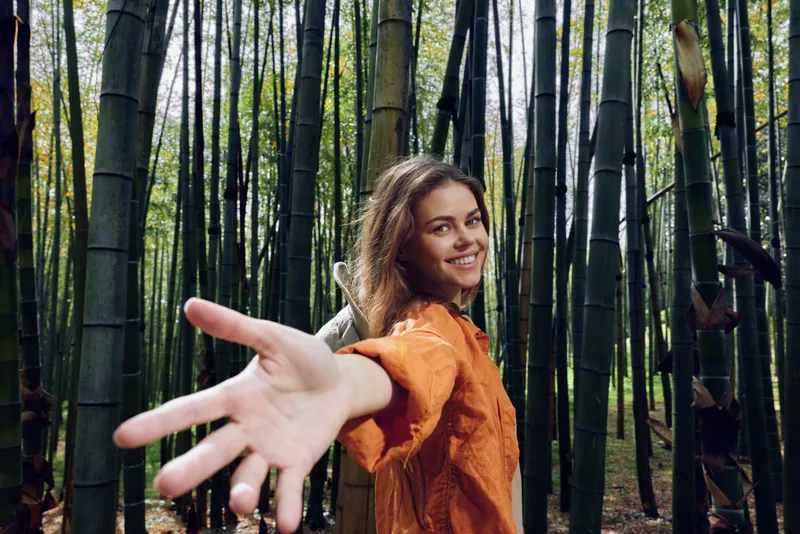 Woman in bamboo forest portrait smiling and reaching hand toward camera, nature scene with tall trees and green foliage, playful outdoor mood and direct eye contact.