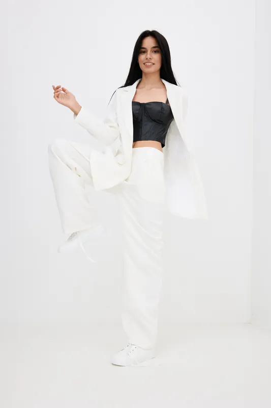 Young woman wearing white suit and black top poses confidently against white background, showcasing modern fashion and casual elegance in minimalistic studio setting.