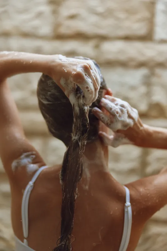Woman washing long hair with shampoo, creating rich foam in shower with warm light and natural stone wall background. Healthy hair care routine concept.