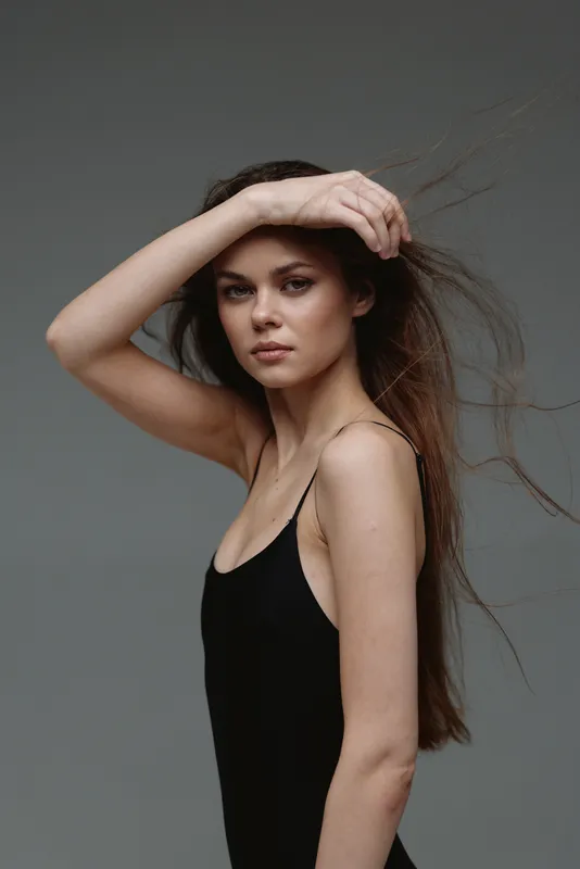serious woman posing with hand on forehead wearing black dress against gray background