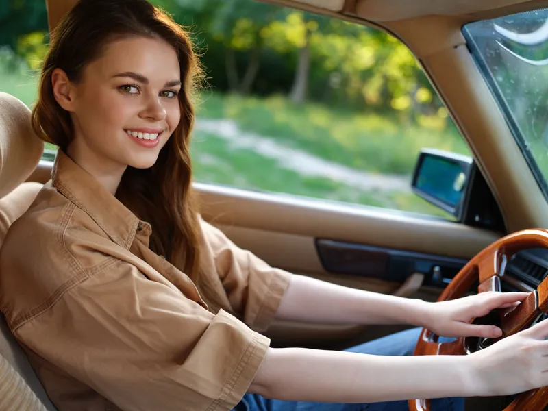 Smiling young woman driving a vintage car in a sunny green landscape, wearing a stylish beige shirt, evoking feelings of freedom and adventure.