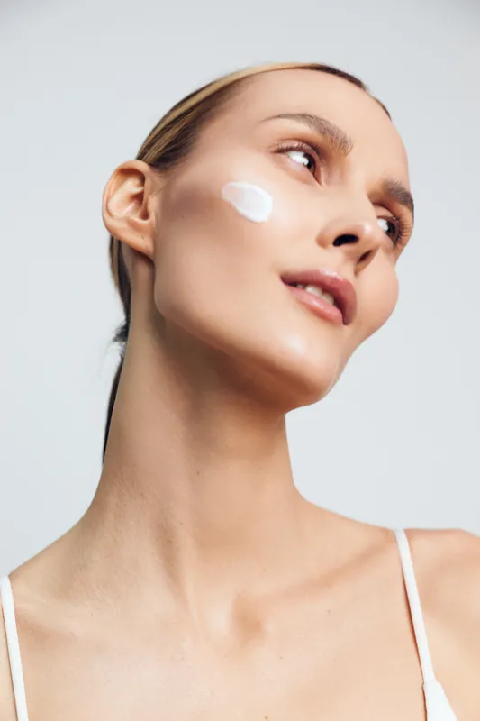 woman skincare beauty face cream cosmetic self care dermatology treatment, portrait of a young female applying moisturizer on her cheek, calm confidence against clean background
