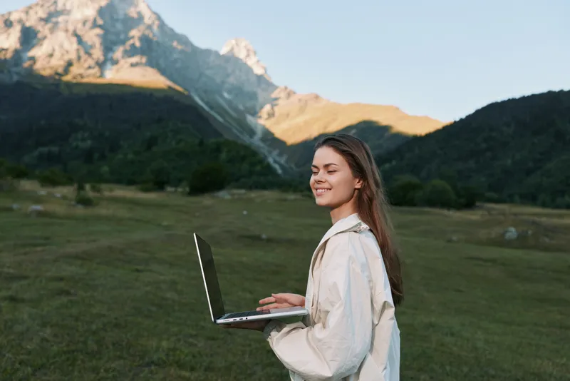 A cheerful woman stands outdoors in a wide grassy field with distant mountains, typing on a laptop. She smiles warmly, exuding calm confidence and outdoor work enthusiasm.