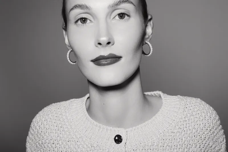 Close-up portrait of a Caucasian woman with fair skin wearing earrings and a textured sweater in black and white studio lighting.