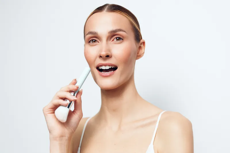 Woman applying skincare serum to face, holding a dropper near the cheek, clean studio background, soft lighting, fresh expression, wellness and selfcare concept, closeup portrait, skincare routine