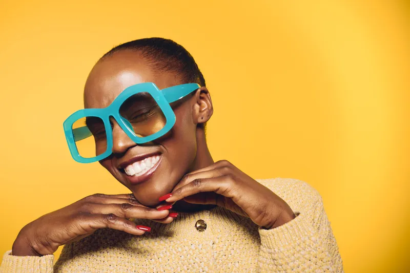 Smiling black woman with dark skin wearing large blue glasses and beige sweater poses happily with hands under chin on yellow background. Concept of joy, fashion, and confident beauty.