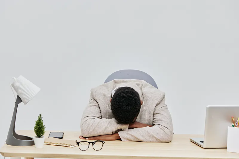 Overworked man resting his head on desk in office environment, exhausted and stressed, surrounded by workplace items like laptop, glasses, potted plant, and notepad, concept of work fatigue and burnou