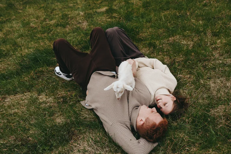A couple lies on the grass in a cozy outdoor setting, sharing a quiet moment, heads close, wrapped in sweaters. A lifestyle scene emphasizing relaxation, affection, and simple leisure together.