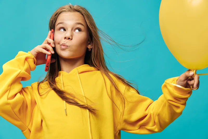 Girl in yellow hoodie holding yellow balloon and talking on red phone with playful expression on blue background. Casual style, communication and fun with colorful contrast and youth.