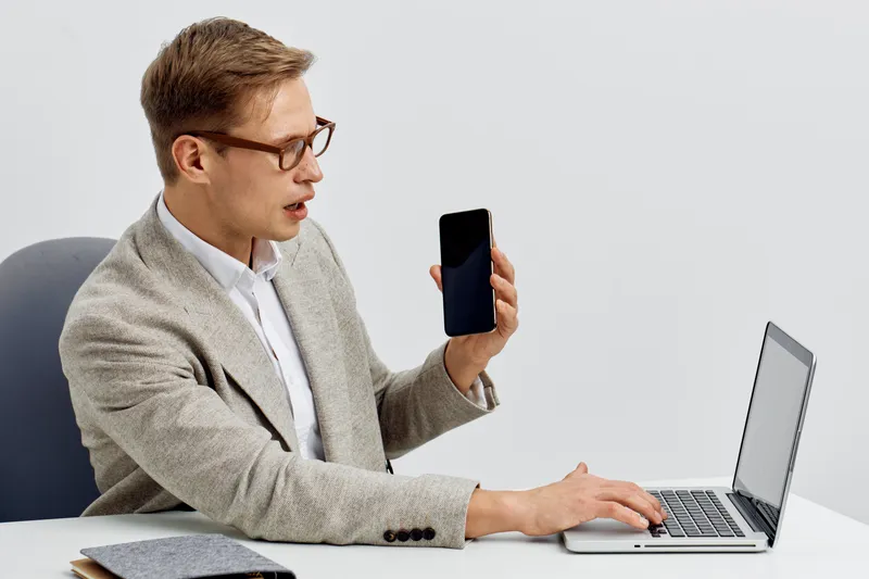 Man using laptop and holding smartphone in modern office, professional workspace, technology, communication, digital devices, male worker, focused, casual business style, trendy environment.