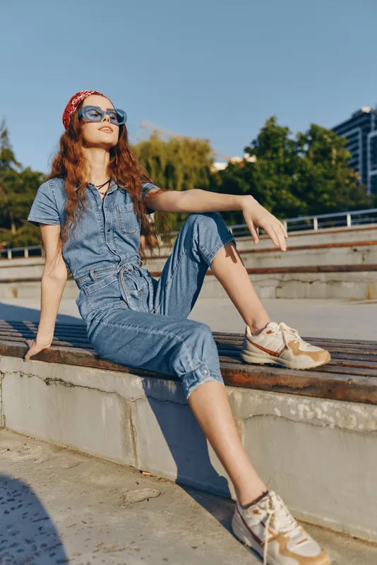 Lifestyle woman in modern boho-western denim street style sitting outdoors with sunglasses and bandana, enjoying sunny day with relaxed confident pose in film color.