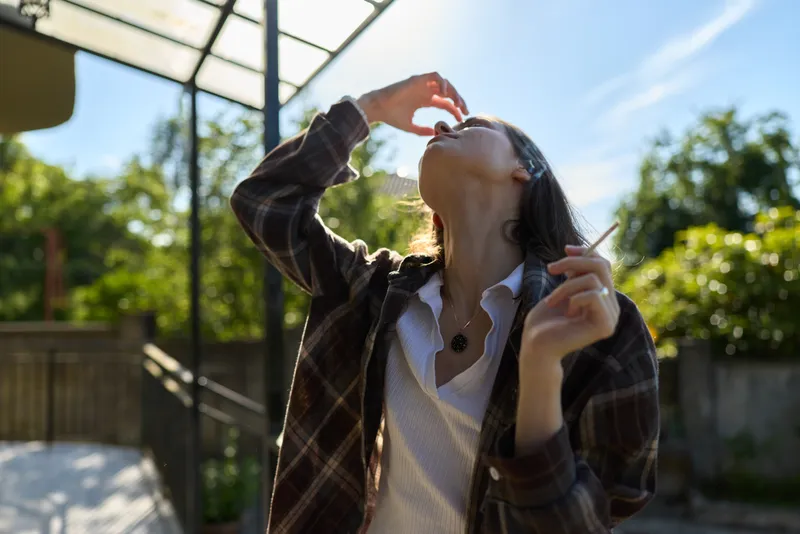 Casual woman enjoys a sunny outdoor moment, drinking from a bottle, wearing a plaid jacket over a light shirt, relaxed lifestyle scene captured in natural daylight