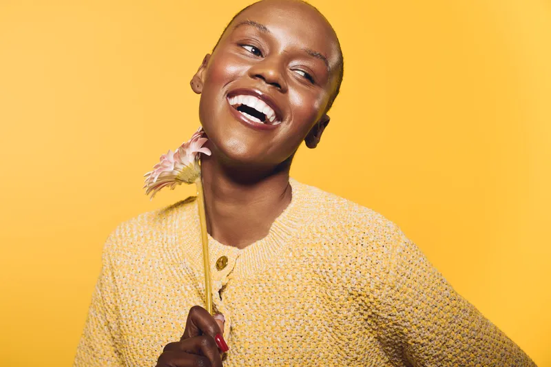 Joyful dark skinned African woman with shaved head smiles holding a flower on yellow background. Concept of happiness, beauty, natural skin, and positive emotions in studio portrait.