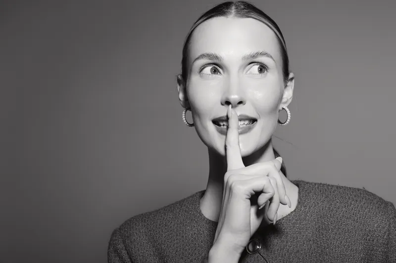 Young white woman with light skin holds finger to lips in silent gesture, looking sideways. Black and white portrait with hoop earrings and casual sweater.