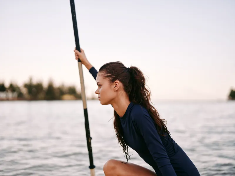 Athletic young woman rowing on calm water with focused expression during outdoor workout in natural setting.