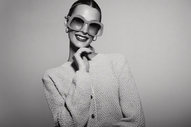Smiling young white woman with pale skin wearing large sunglasses and textured jacket posing against grey background.