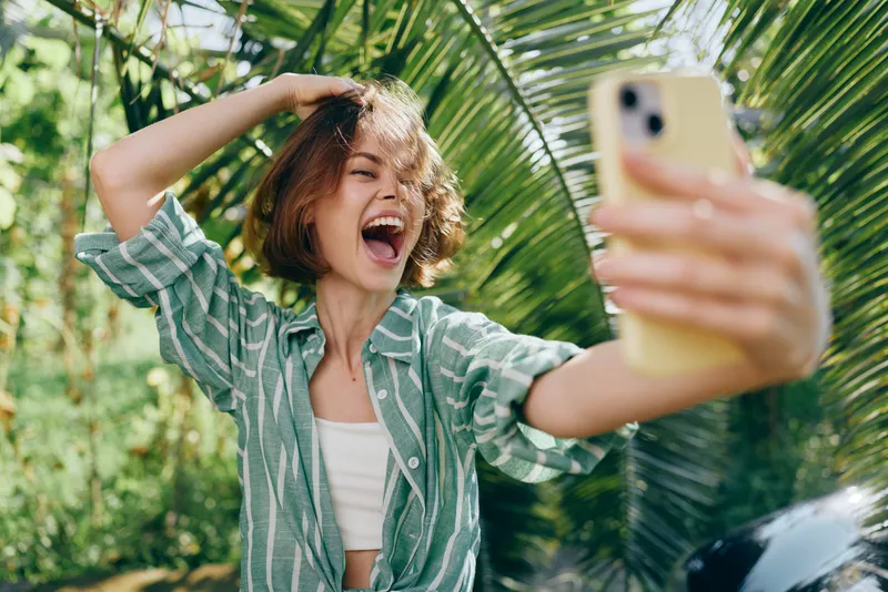 Young woman taking a vibrant selfie outdoors in a sunny tropical setting, smiling with excitement.