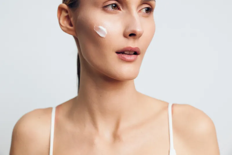 Young woman with clear skin applying cream on face, natural beauty concept, soft lighting, white background, close-up portrait, skincare routine, calm expression.