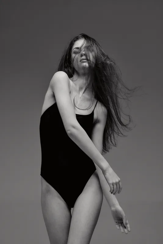 Black and white graceful woman in black swimsuit with flowing hair posing in studio, expressing movement and elegance.