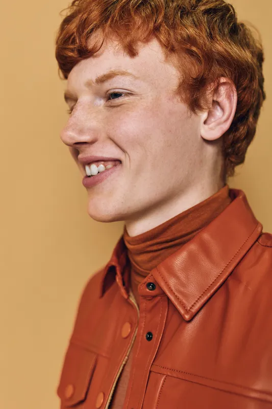 Young redhead man smiling in profile wearing a brown leather jacket and turtleneck against a beige background with soft natural lighting and casual style.