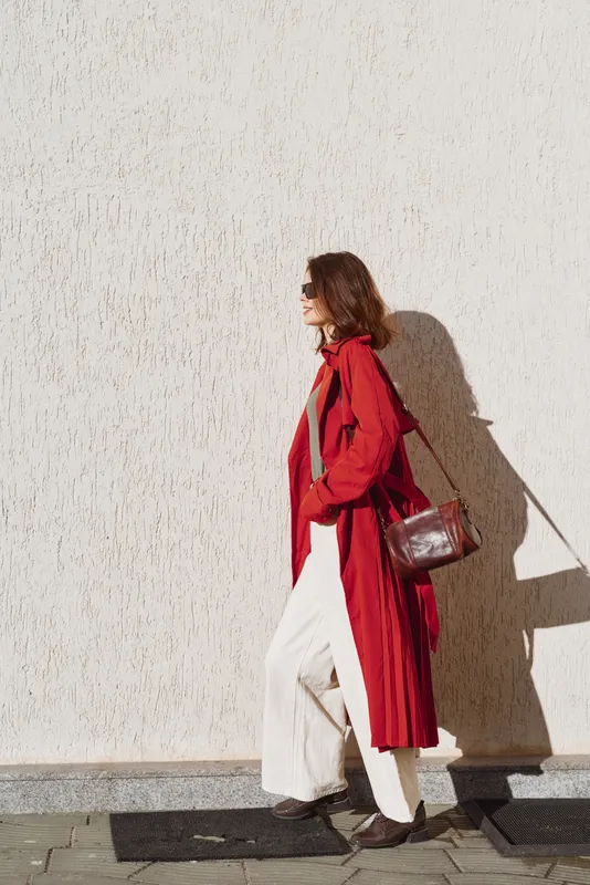 A woman in a long red coat and white pants walks along a textured beige wall in bright sunlight, carrying a brown leather bag. Casual urban style with strong shadow on the wall.