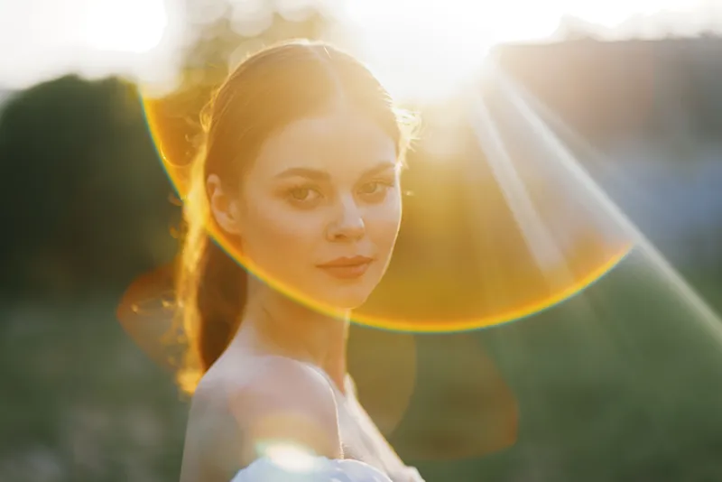 Young woman in a sunlit field, wearing a white dress, radiating warmth and serenity with a soft smile against a vibrant sunset background