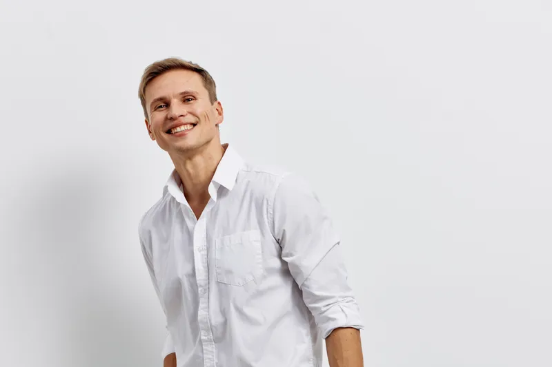 Smiling young man with light brown hair wearing a white shirt looks joyful against a plain white background. Studio portrait capturing happiness, positive attitude, casual style, and approachable deme