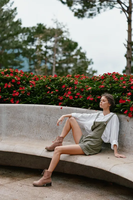 Young model in stylish green overalls and white blouse, sitting gracefully in a park with blooming red flowers, conveying a sense of calm and connection to nature
