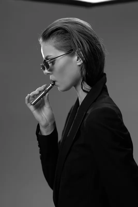 Black and white portrait of a stylish woman vaping with sunglasses and sleek hair in a minimalistic setting.