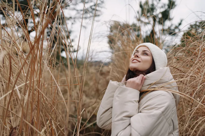 Woman in a light coat and beanie stands among tall grasses, looking upward with a thoughtful expression, outdoor fashion and natural scenery for a seasonal mood