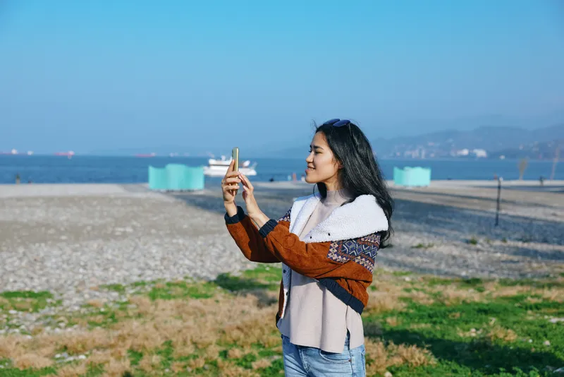 Beach scene with a woman using a smartphone to capture a moment, sunny weather, casual jacket, sea horizon, distant boats, coastal grass, relaxed mood, outdoor photography memory