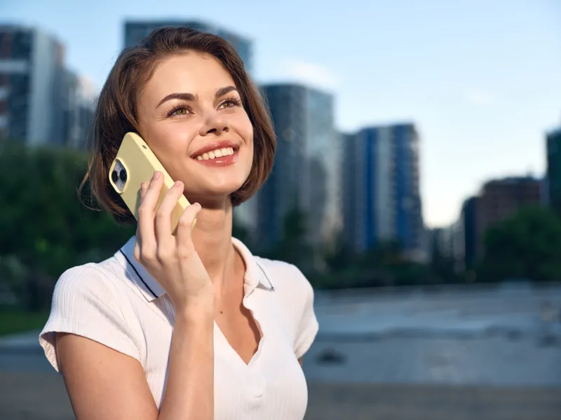A cheerful woman speaks on a mobile phone in an outdoor urban setting. She wears a light blouse and smiles brightly, projecting confidence, optimism, and professional poise.