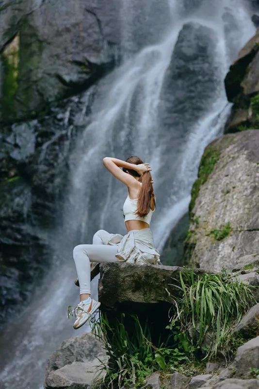 Young woman sitting on rock near waterfall in casual sportswear, enjoying nature, relaxed posture, outdoor adventure, green moss background, peaceful atmosphere.