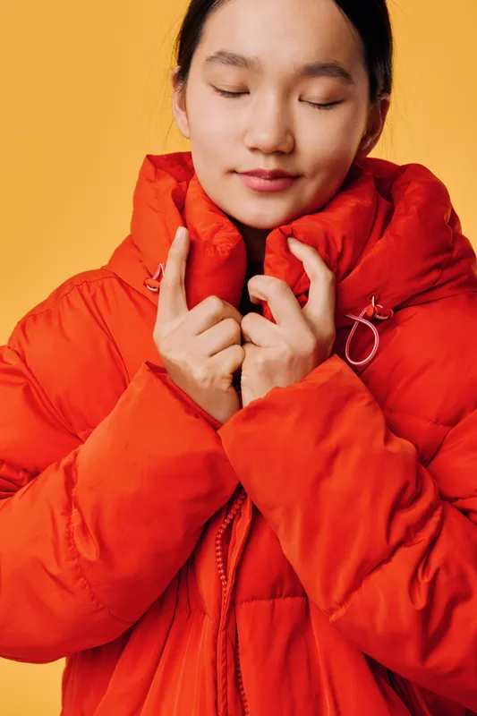 Woman in red puffer jacket with closed eyes holding collar for warmth and comfort against yellow background. Portrait of young adult with cozy winter clothing and peaceful expression.