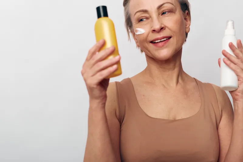 Mature woman in beige top applying white cream on cheek holds two cosmetic bottles. Studio shot on light background. Concept of skincare choices, beauty routine, and self-care for aging skin.
