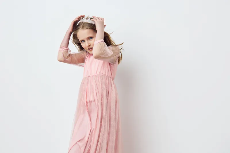 Young girl in a soft pink dress, playfully posing and smiling with a crown, showcasing emotions of joy and innocence in a minimalistic studio background