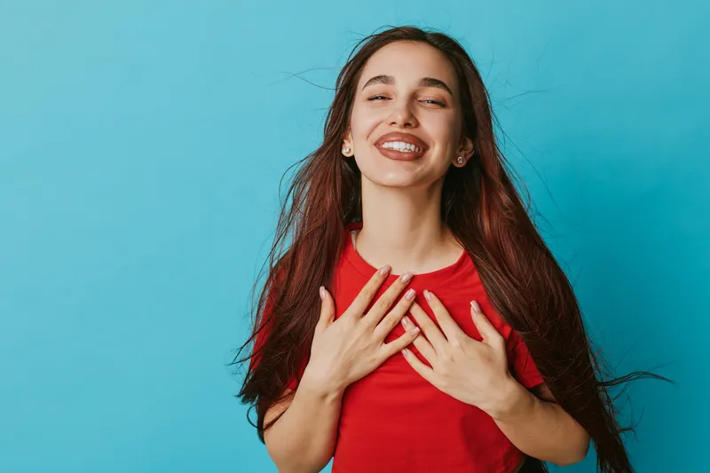 Young woman with long hair and braces smiling with closed eyes and hands on chest against blue background. Happiness, gratitude and positive emotion in casual red shirt portrait.