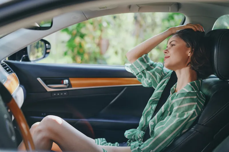 A woman sits in a car seat, gazing through the window with her hand on her forehead, enjoying a sunny, relaxed drive.