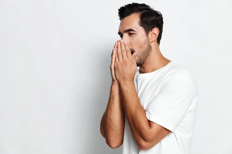 Young caucasian man showing surprise, covering mouth with hands, wearing a casual white t-shirt, isolated on plain solid gray background. Emotion expression concept.