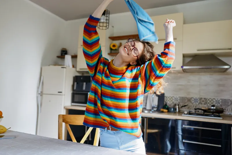 Woman in a home kitchen enjoys a playful moment, tossing a blue cloth while wearing a colorful striped sweater, bright lighting and casual lifestyle atmosphere.