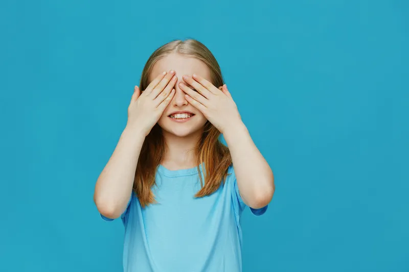 Smiling girl covering her eyes with hands against a bright blue background, displaying joy and innocence in a playful manner ideal for children s themes
