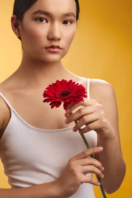 asian woman holds a red flower against a warm yellow backdrop, presenting a calm, thoughtful mood with soft lighting, simple clothing, and graceful poise.