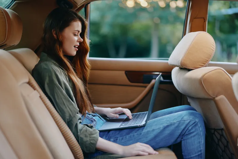 Young woman working on a laptop in a car interior, wearing casual clothing and expressing a focused emotion, with a natural green background.