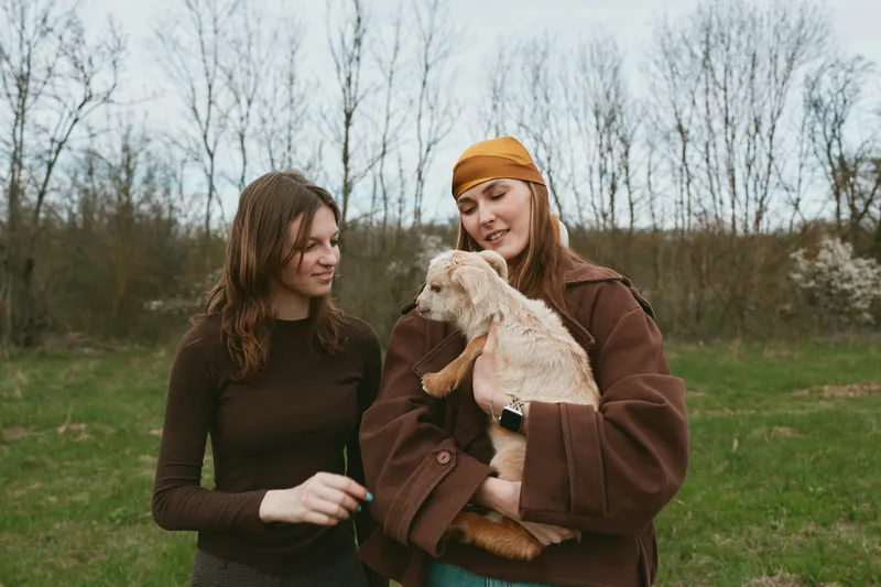 Two young women enjoy a casual moment outdoors as one holds a small goat while the other smiles, surrounded by bare trees and green grass in a relaxed lifestyle scene.