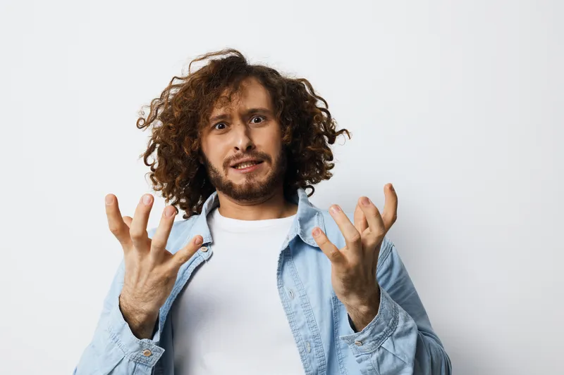 Young man expressing frustration with messy curly hair, wearing a denim jacket, against a plain white background, reflecting emotions of disbelief and exasperation